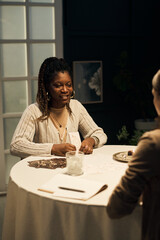 Black young woman reading tarot cards for Caucasian middle aged woman at round table, both sitting...