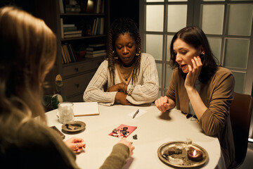 Two young women, one Black and one Caucasian, sitting at round table, receiving tarot card reading...