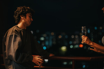 Two young adults engage in a relaxed conversation on a balcony at night, holding drinks. The city...