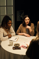 Caucasian young woman and Black girlfriend sitting at round table listening to female psychic,...
