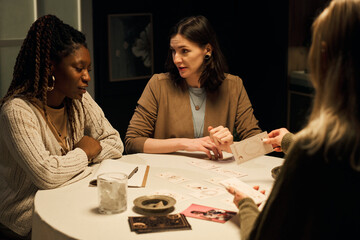 Caucasian and Black young women sitting at round table discussing card predictions and meanings while female psychic is laying out tarot during fortune reading