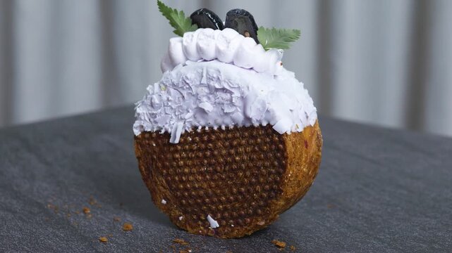 Half of a cronut pastry standing upright, featuring a textured, caramelized base and topped with lavender icing, whipped cream, fresh mint leaves, and blackberries on a dark surface