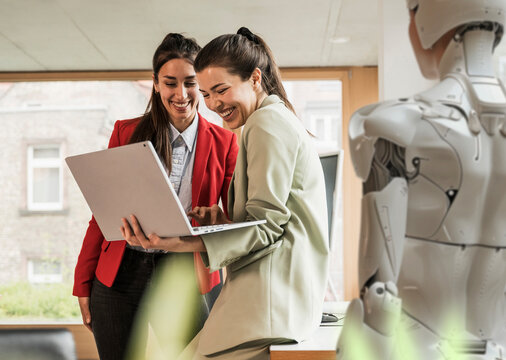 Cheerful businesswomen using laptop and standing with colleague in office