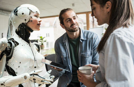 Businesswoman talking with colleague holding tablet PC and standing near cyborg in office