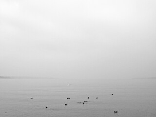 A moody, monochromatic view of a lake on a foggy, overcast day. The surface of the water is calm, and a line of small, dark boats is barely visible, conveying a sense of solitude and mystery.