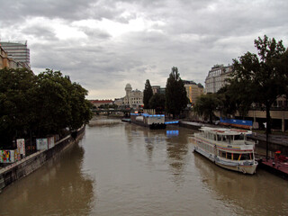 A passenger boat on the Danube Canal (Donaukanal) in Vienna, Austria, on a cloudy day. The urban cityscape with historic and modern buildings lines the riverbanks.