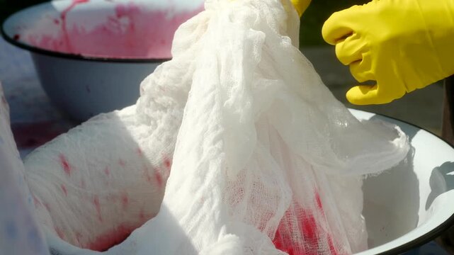 Experienced winemaker wearing yellow protective gloves carefully filtering freshly squeezed cherry juice through cheesecloth in large metal basin during sunny summer day