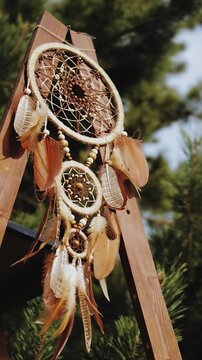 Vertical video. Dreamcatcher with brown feathers hanging on a wooden board. Wedding decor for a boho wedding. Feathers swaying in the wind against the background of pine trees.