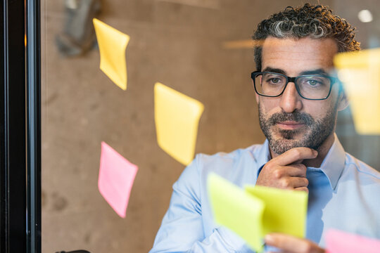 Thoughtful businessman sticking adhesive note on glass at co working space
