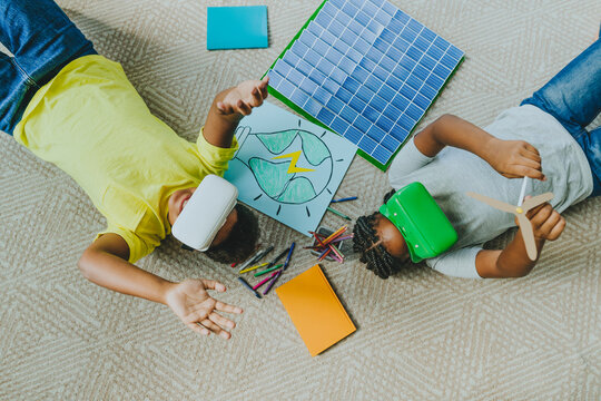 Siblings wearing virtual reality simulators and lying near solar energy school project on carpet