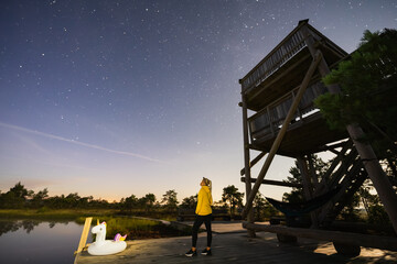 A woman in a yellow hoodie is stargazing at a magical night sky by a wooden observation tower in...