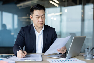 Serious young Asian man in a suit working in the office on documents, studying papers and writing down data