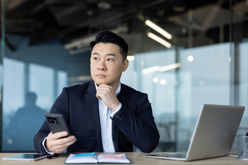 Fototapeta premium Serious young Asian male businessman sitting in the office at his desk, holding a phone in his hand and looking thoughtfully out the window