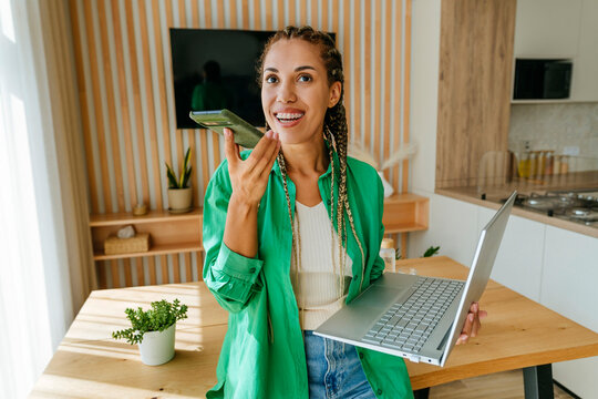 Happy freelancer sending voicemail through smart phone and standing with laptop at home