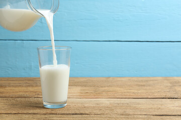 Pouring fresh milk from jug into glass at light wooden table in kitchen, closeup