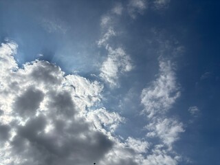 Bright blue sky with a smooth spread of white clouds, sunlight is seen spreading softly from behind the cloud layer in the middle, creating a calm and peaceful atmosphere.