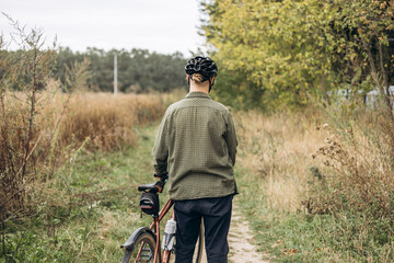 Back view of a man cyclist in casual clothes and helmet walking near a field.
