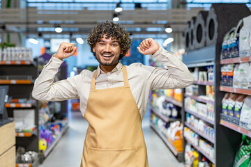 A cheerful retail employee wearing an apron celebrates while standing in a bright store aisle. The...