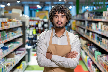Fototapeta premium Supermarket worker stands with arms crossed in a vibrant grocery store, showcasing professionalism and pride in a retail environment.