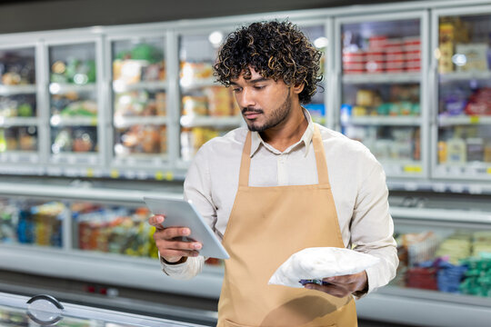 Young employee in a supermarket wearing an apron, analyzing a tablet and holding a frozen item in the frozen foods aisle. The setting emphasizes the modern technology used in the retail sector. - Powered by Adobe
