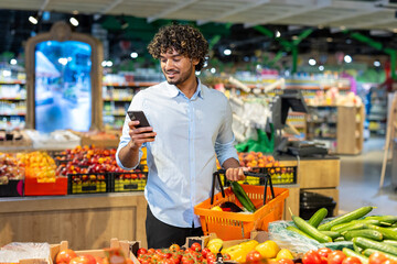 A young man browsing his smartphone while selecting fresh vegetables in a supermarket. He holds an orange basket with groceries as he explores the store's produce section amidst colorful displays.