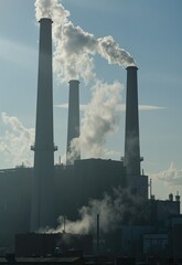 A large industrial complex with multiple tall chimneys emitting white smoke into a clear blue sky, illustrating the presence of pollution and power generation