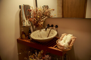 Rustic bathroom sink with stone basin, wall-mounted faucet, decorative dried flowers, and rolled towels on wooden countertop