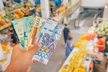Hand holding Kazakh tenge banknotes at a blurred fruit and vegetable market in Almaty, Kazakhstan