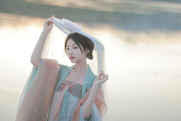 Young woman in traditional attire holding a hat by the water during sunrise.
