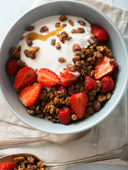 A bowl of granola, yogurt, and sliced strawberries is pictured. The bowl is light blue and the food...