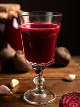 A glass of vibrant red beet juice, with raw beets and garlic cloves scattered around a wooden surface. A jar of what appears to be beets is in the background.