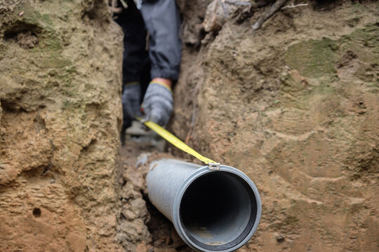 Construction worker in safety gear measuring and aligning a plastic drainage pipe in a trench during septic system installation at a private house, ensuring proper slope and connection for local sewag