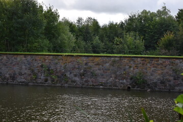 Stone wall along river surrounded by trees under cloudy sky  