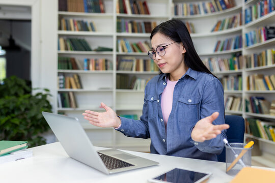 A woman sitting at a desk in a library, gesturing frustratedly while working on her laptop, surrounded by books, expressing problem-solving or challenges in a study or professional context.