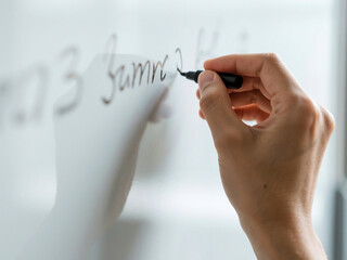 Close-up of a person’s hand writing on a whiteboard using a black marker pen, ideal for illustrating concepts such as brainstorming, teaching, business presentations, planning, or corporate training