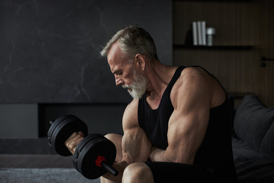 Middle aged Caucasian man exercising with dumbbell, sitting on sofa, focusing on strength training, showing muscular arms and concentrated facial expression, looking downward