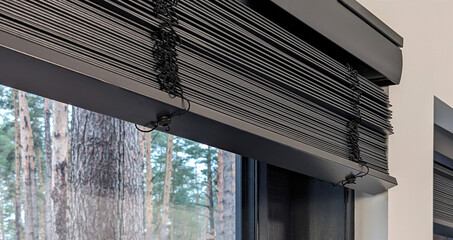 Motorized wooden blinds are folded up on a large window. Black wood shutters, close-up photo. Curtains are installed in the window opening. Outside the window is a pine forest.