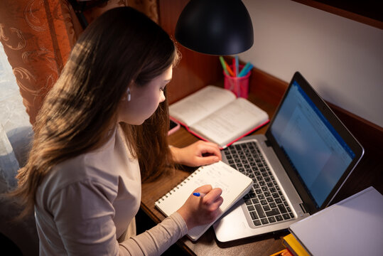 Student focused on homework while wearing headphones. A young woman sits at a desk with a laptop, writing in a notebook, focused on her task while wearing headphones
