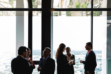 Group of diverse professionals talk casually near tall windows in a high-rise office. Relaxed...