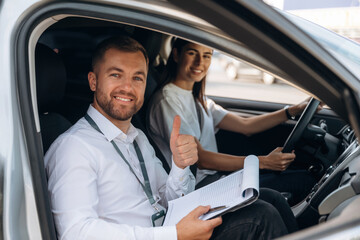 Gesture of thumb up, smiling. Woman is with instructor in car, driving school concept