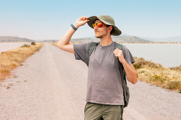 Young male tourist adjusting his hat while strolling along a coastal road, enjoying the sunny...
