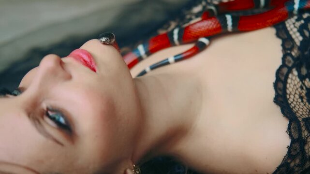 Gothic fantasy woman dark queen of snakes and poisons. Gothic Girl vampire Beauty princess is lying down, hugs red snake sinaloa. Flowing hair, black dress. Royal milk snake crawls on chest. video 4k