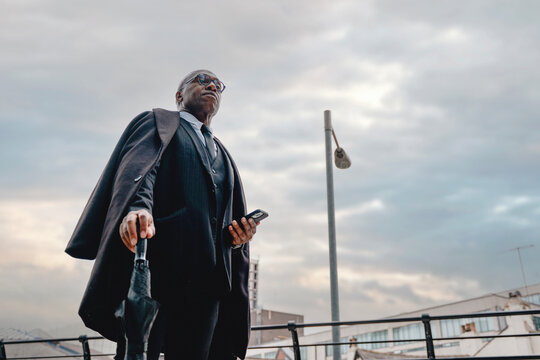 Businessman stands confidently with umbrella in hand against cloudy sky while checking phone near urban setting during late afternoon