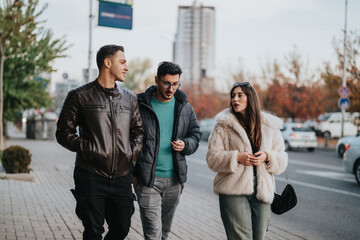 Fototapeta premium Young friends enjoy a casual walk in the city during autumn, discussing and enjoying the outdoors surrounded by urban and natural settings. The scene captures social interaction and warm friendship.