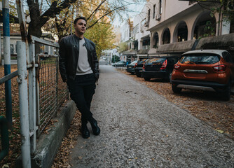 A man casually leaning by a fence along a leafy street during autumn, characterized by parked cars and vintage architecture. The scene reflects leisure and a serene city atmosphere.