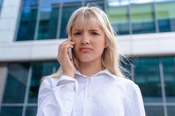 Young woman looks concerned while talking on a smartphone outside a modern building during the...