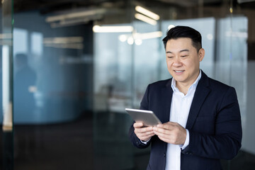 Fototapeta premium Asian young businessman in a suit standing in the office lobby and using a tablet