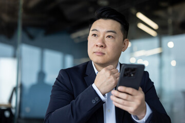 Close-up photo of a young Asian man in a suit sitting at his workplace with a phone in his hand and...
