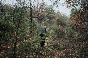 A person walking through a calm woodland filled with green and brown foliage under a cloudy sky,...