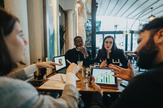 A group of multiracial business colleagues engaged in a collaborative meeting, reviewing financial charts and business strategies on a digital device in a modern cafe setting.